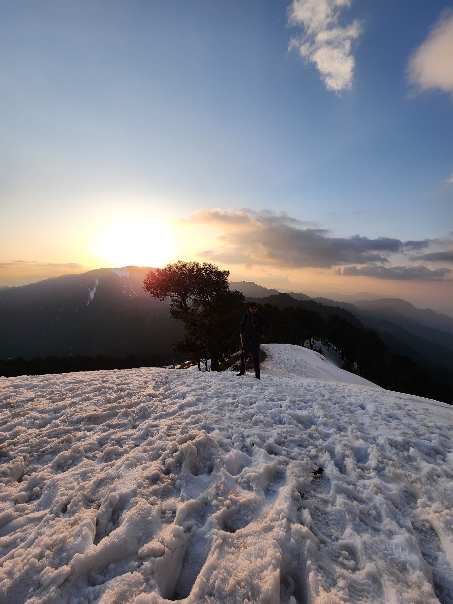 Person standing on a snowy slope overlooking mountains during sunrise