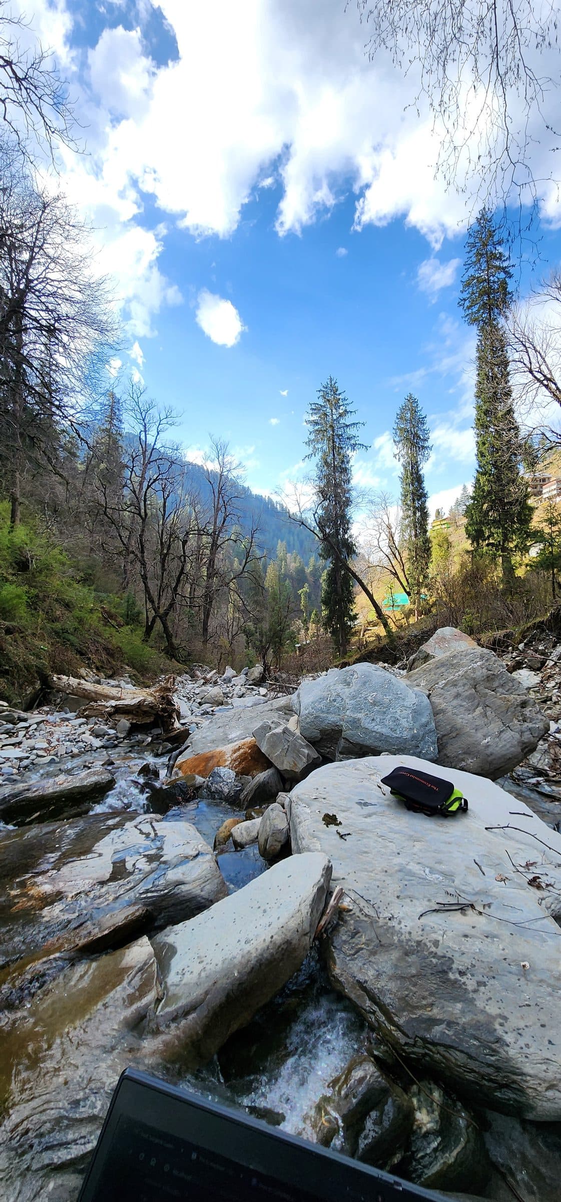 A rocky stream flows through the Shoja Valley in Himachal Pradesh, India, with a laptop resting on a stone.