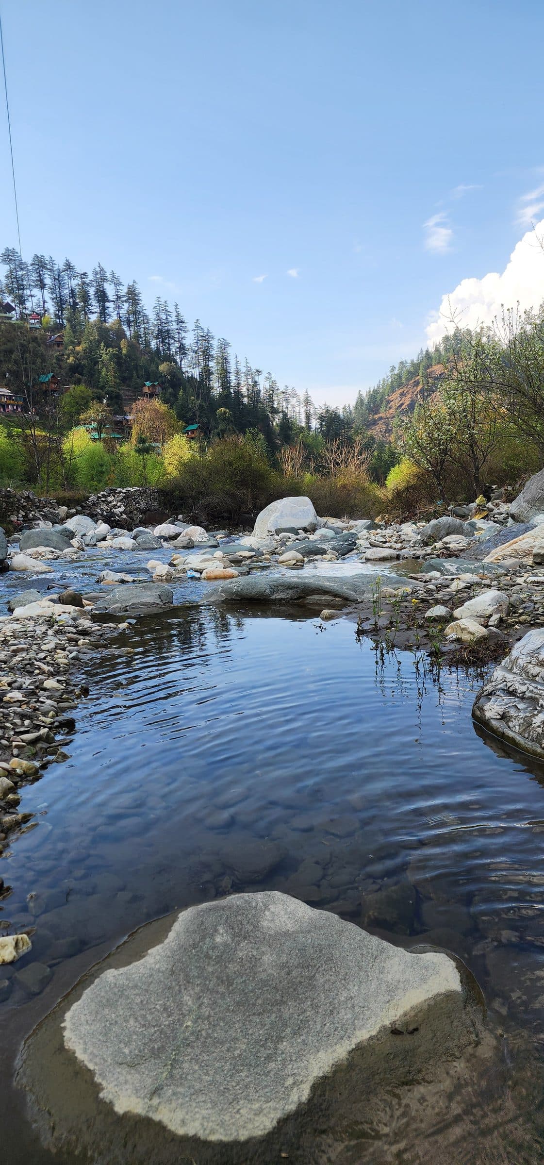 A serene river scene in Jibhi Valley, Himachal Pradesh, India, featuring smooth grey rocks and shallow water.