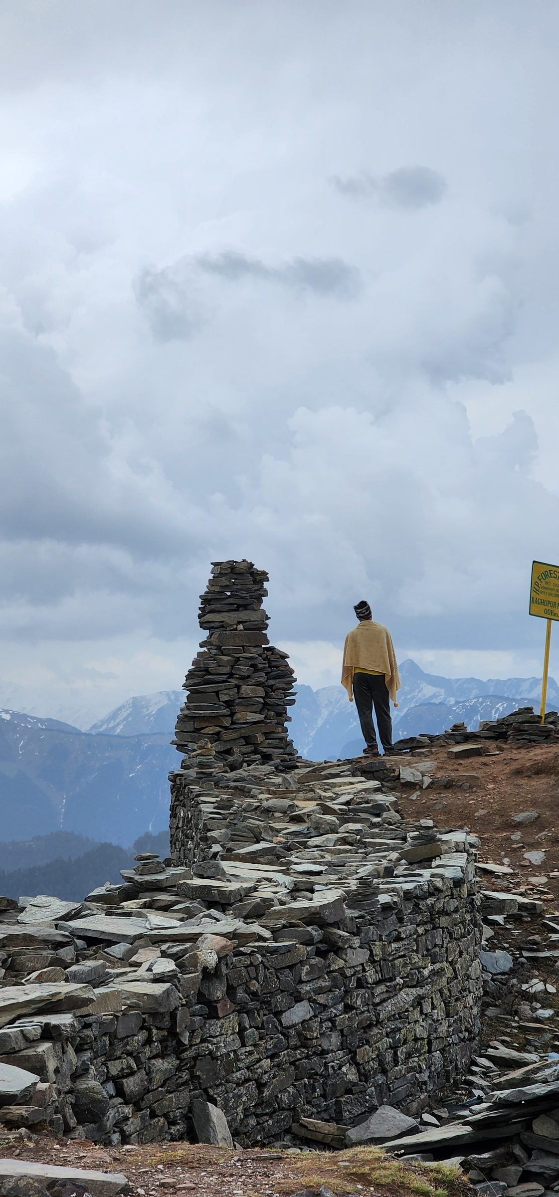 A person stands near a large stack of grey stones overlooking the mountain valley.