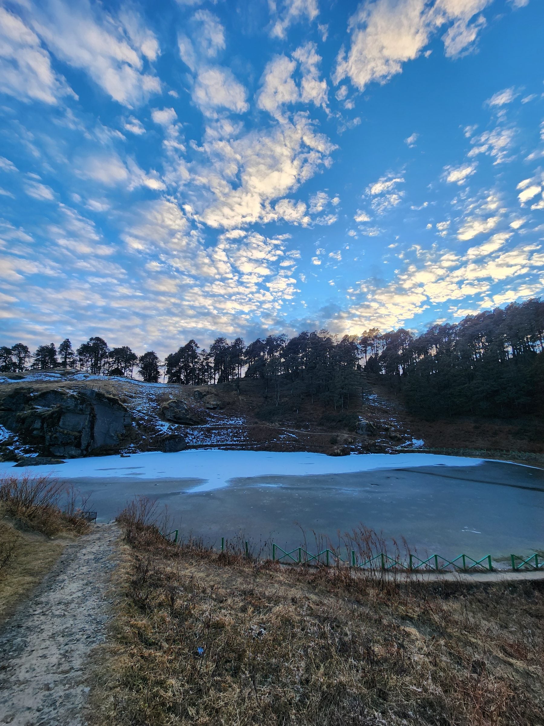 A tranquil frozen lake reflects the winter sky above the snow-dusted hills of Jibhi Valley, Himachal Pradesh.