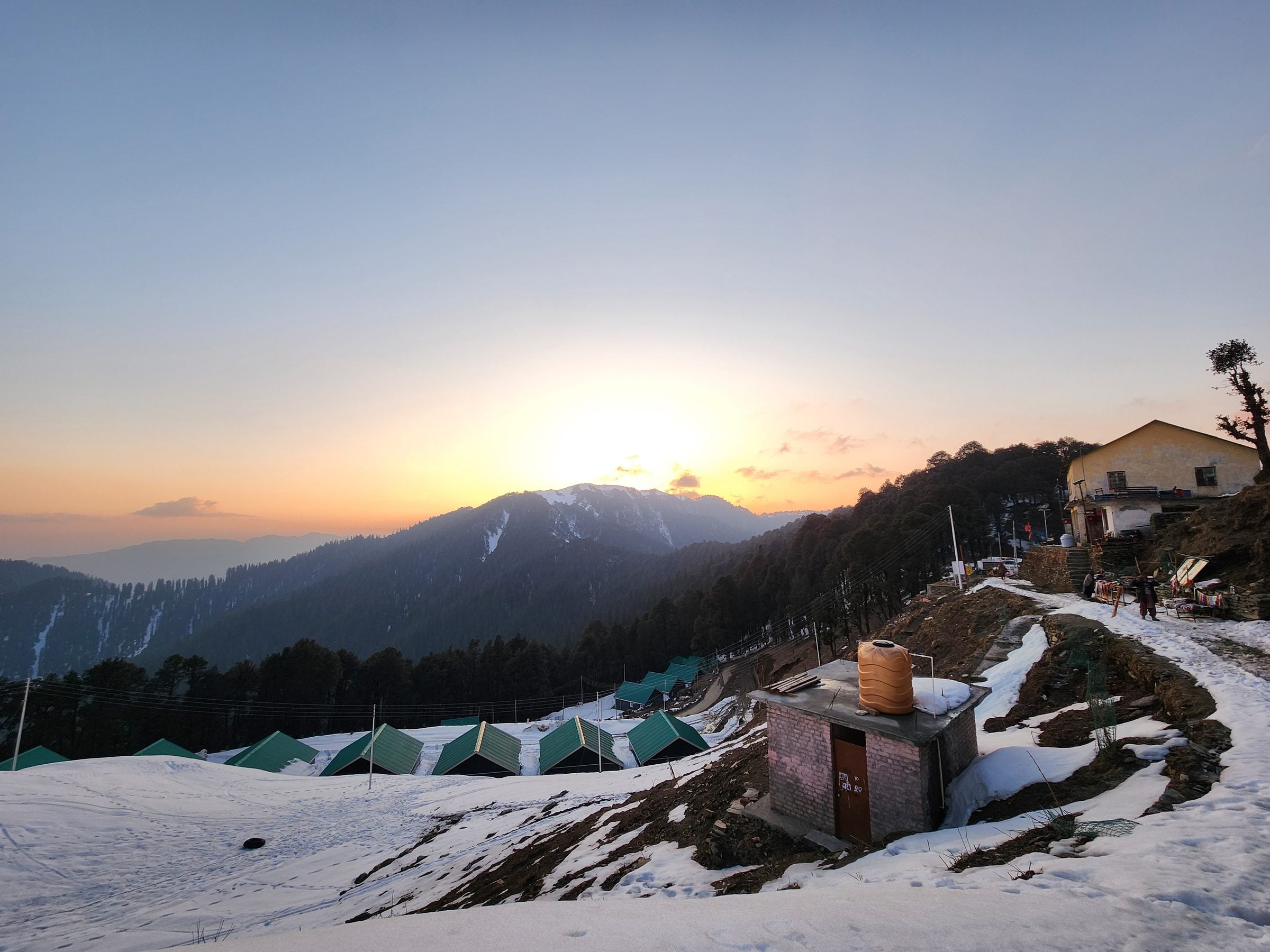 A serene view of a snow-covered hillside in Shoja Valley, Himachal Pradesh, India with a wooden pathway and distant trees.