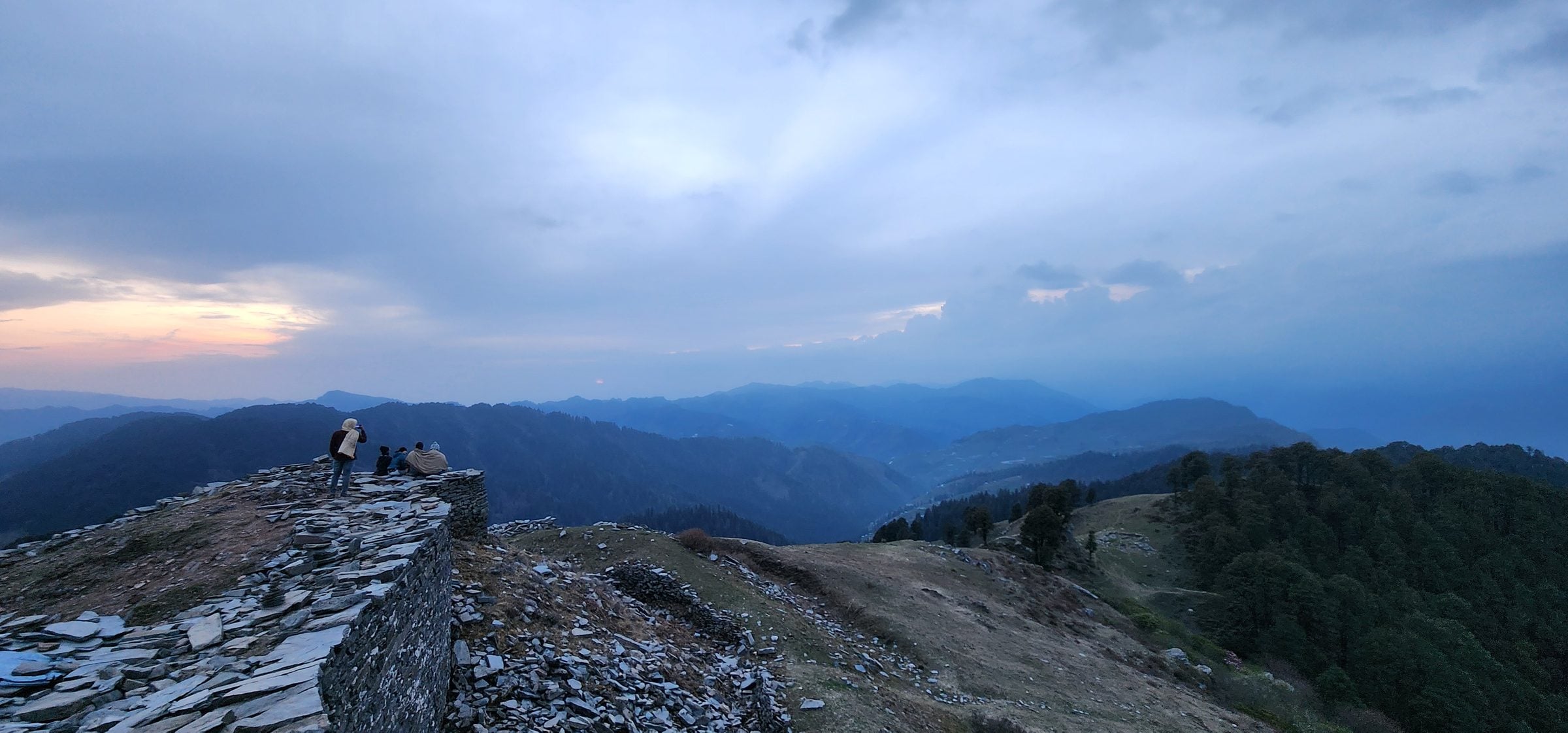 A lone figure enjoys the breathtaking sunset view overlooking the rocky terrain of Jibhi Valley, Himachal Pradesh.