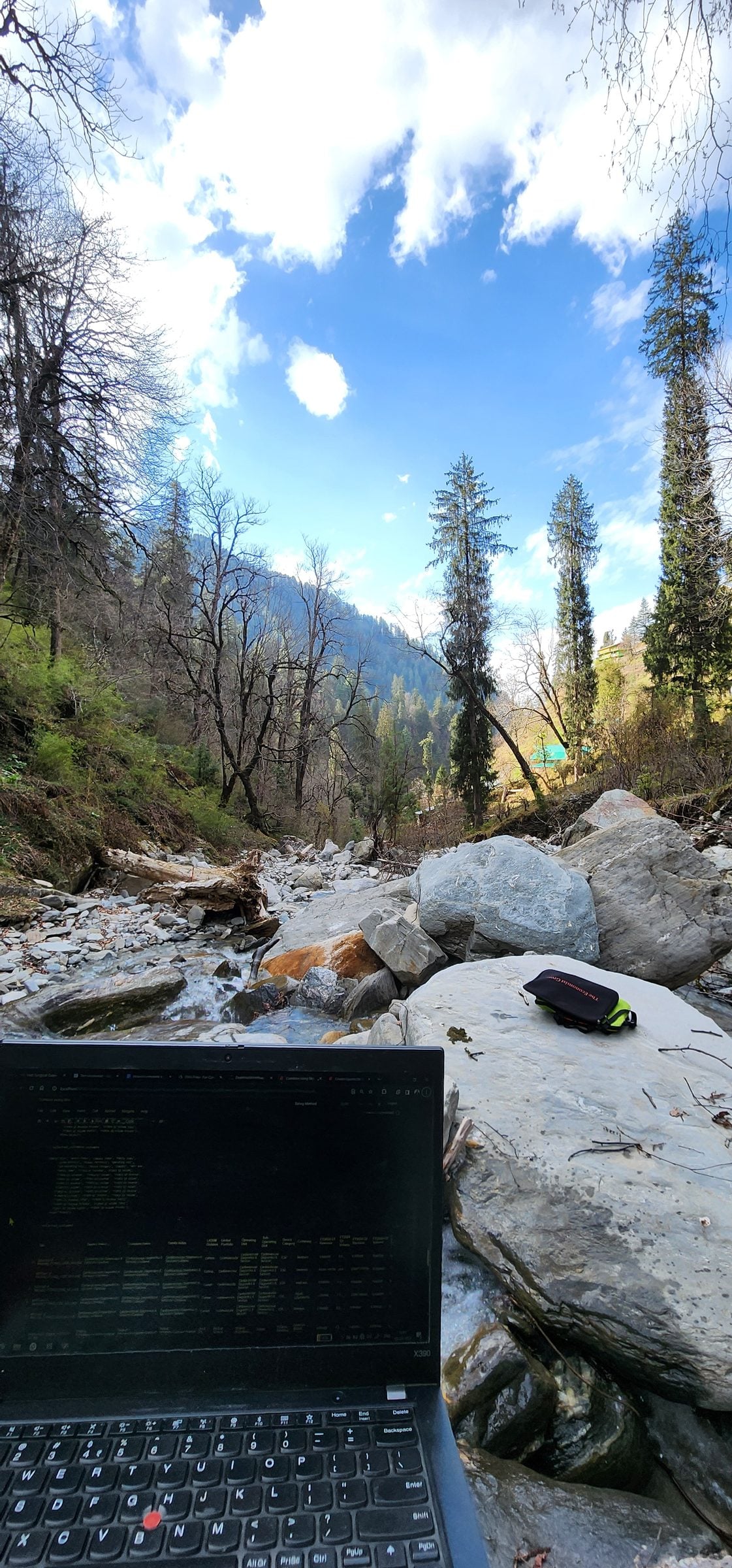 A laptop rests on a rocky riverbank in Jibhi Valley, Himachal Pradesh, India, overlooking a serene mountain stream.