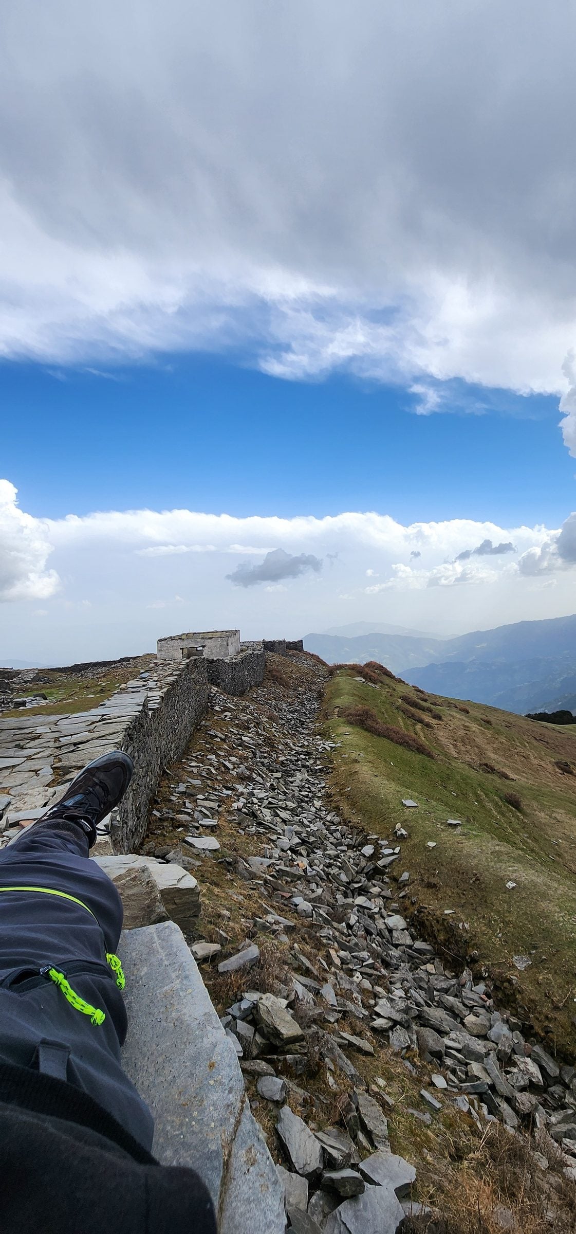 Serenity on a Jibhi Valley Ridge A person sits on a weathered stone wall overlooking a vast, cloudy mountain valley in Jibhi, Himachal Pradesh, India.